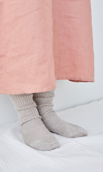 Plümo- Person wearing Linen Rib Socks and a long pink skirt, standing on a white textured surface against a white wall.