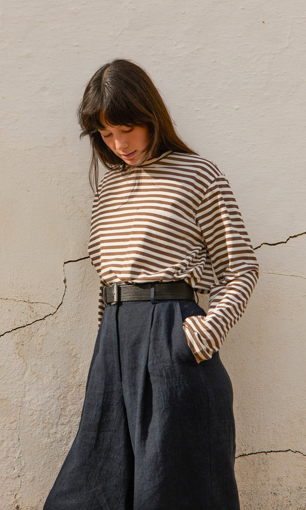 Plümo- Woman in striped long-sleeve shirt and Lucien Shorts - Navy stands against a cracked, off-white wall, looking down.
