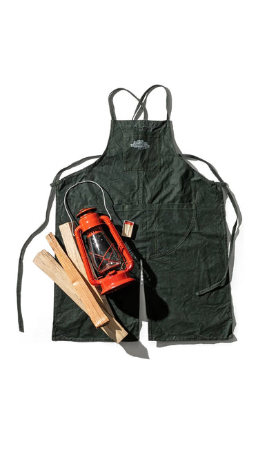 Plümo- Lumberjack Apron in dark green with two front pockets, a red lantern, wooden sticks, and matchbox on white background.