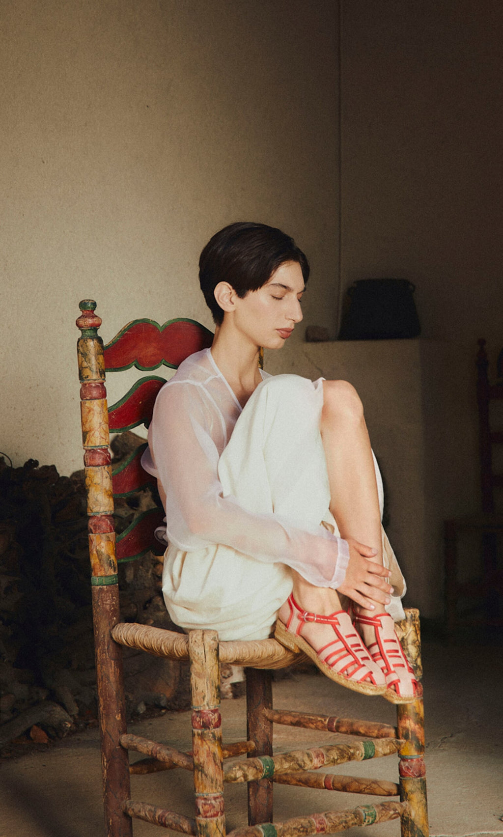 Plümo- Barefoot in a sheer white outfit, they sit by their Cordoba Espadrilles on a rustic chair, gazing down peacefully.