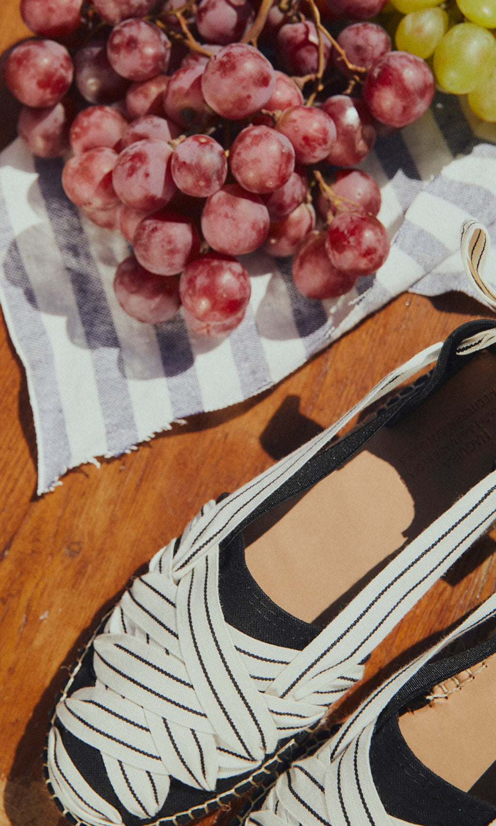 Plümo- Ribbon Espadrilles with jute wedge placed beside grapes on a striped cloth over a wooden surface.