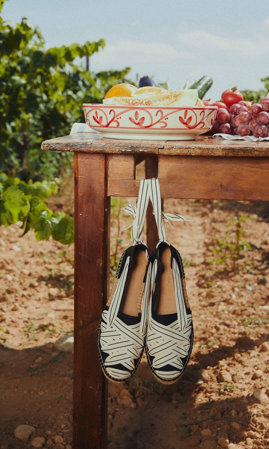 Plümo- Ribbon Espadrilles with striped design and jute wedge hang from a wooden table near a vineyard.