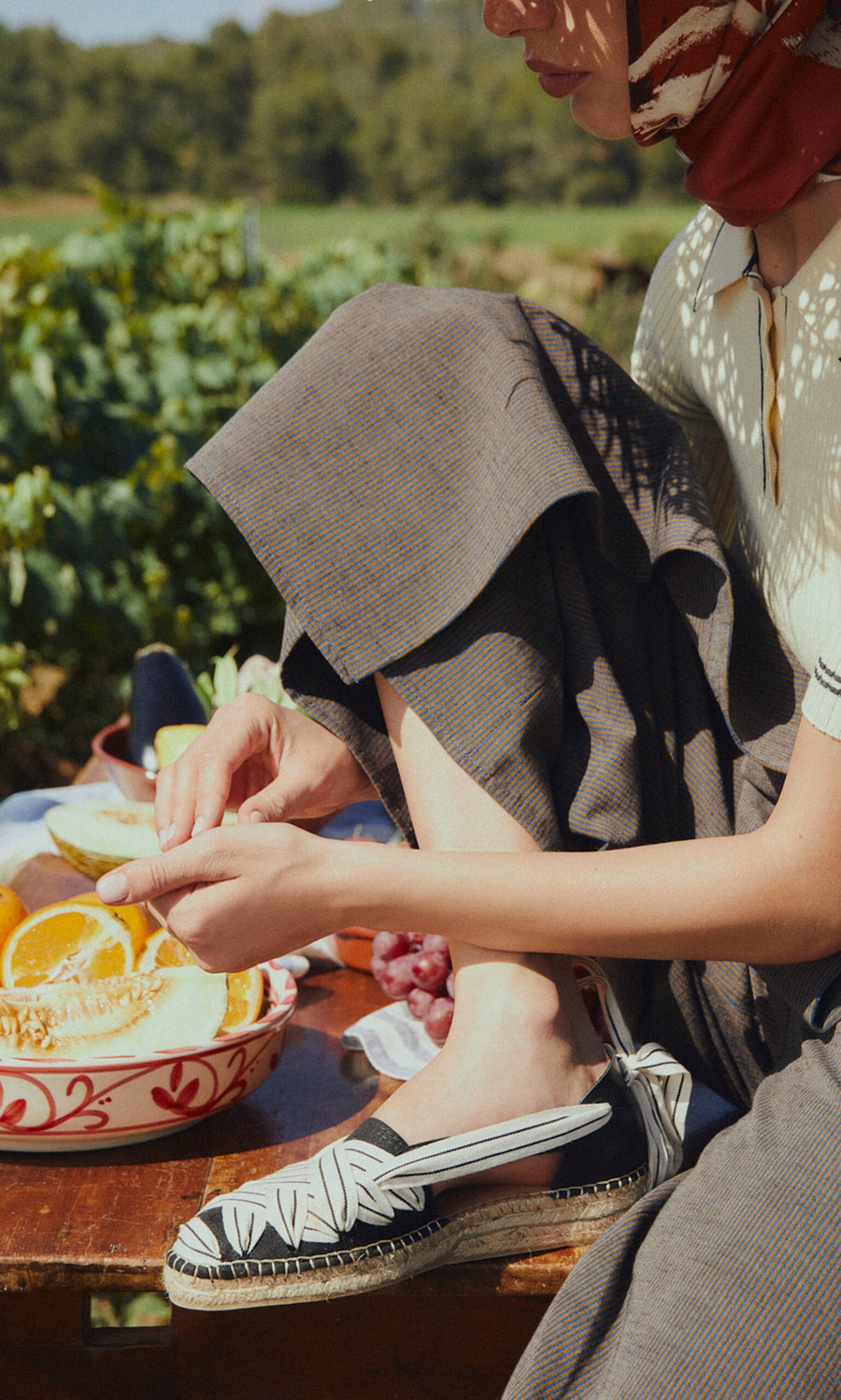 Plümo- Woman outdoors peeling citrus, wearing Ribbon Espadrilles with a jute wedge, a scarf, and a brown skirt.