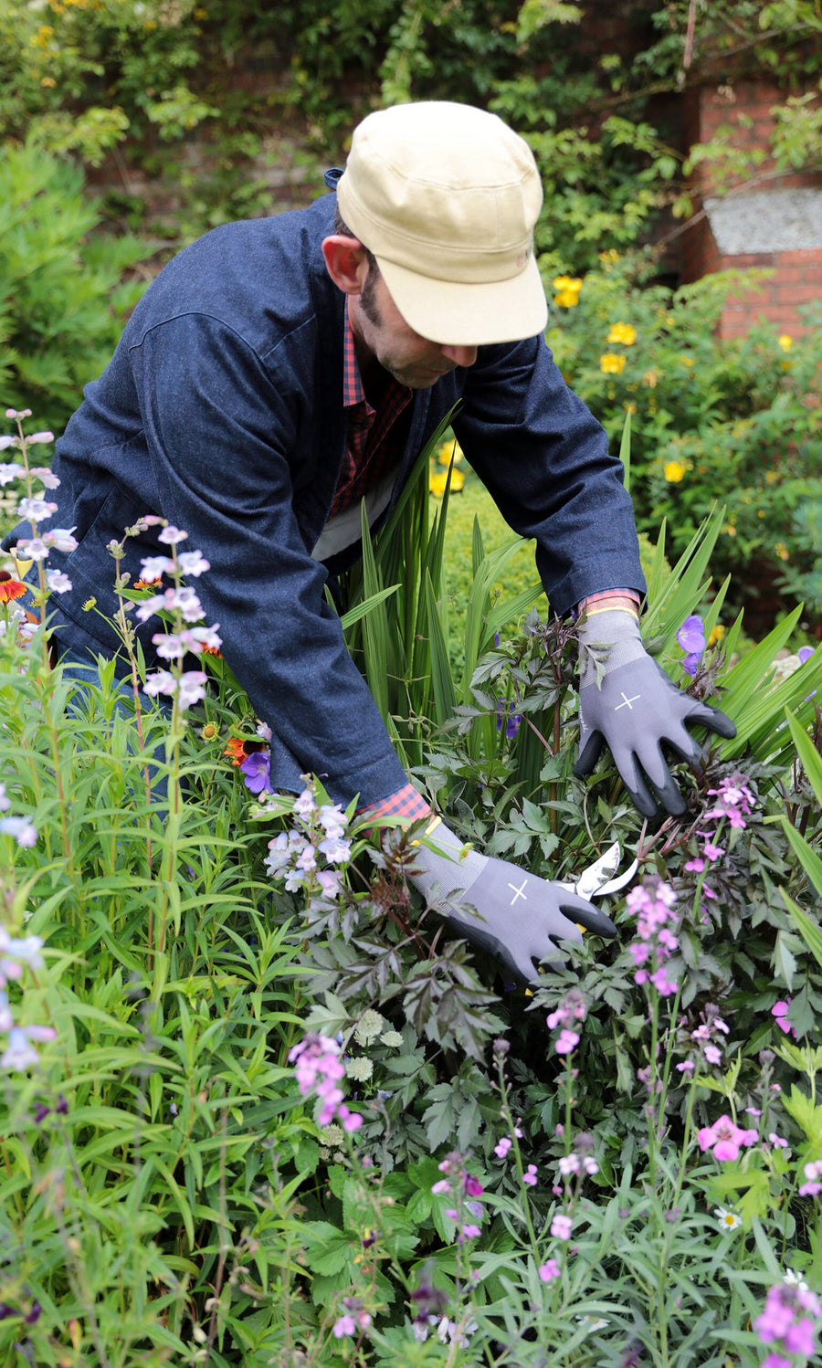 Plümo- Person wearing Niwaki gardening gloves trims purple flowers among lush greenery and vibrant garden blooms.