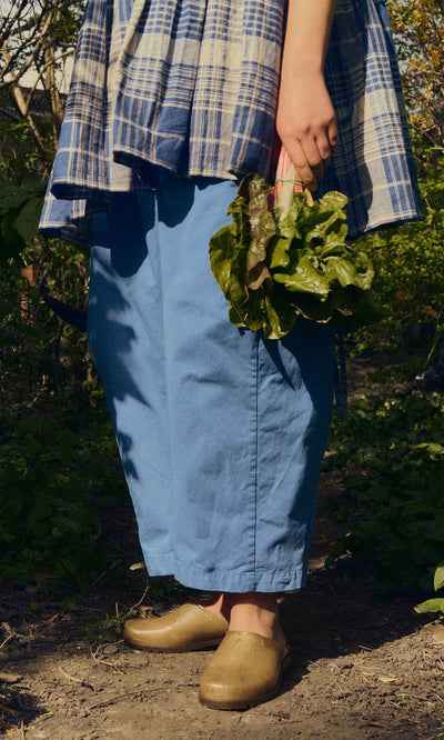 Plümo- Wearing Opana Mules, a person in blue pants and plaid shirt holds leafy greens on a sunlit dirt path.
