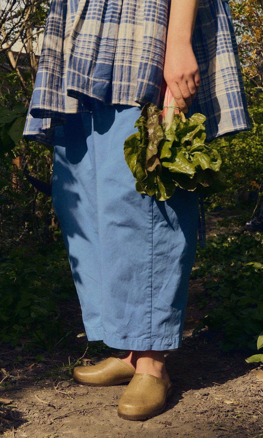 Plümo- Wearing Opana Mules, a person in blue pants and plaid shirt holds leafy greens on a sunlit dirt path.