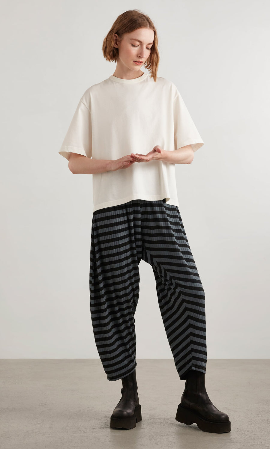 Plümo- A woman stands indoors on a gray floor wearing Oyster Catcher Pants - Grey Stripe and black platform boots.
