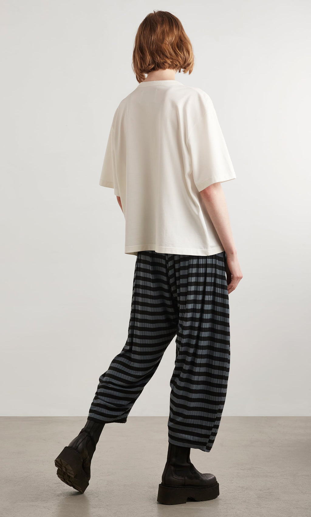 Plümo- Person with short hair in a loose white top, Oyster Catcher Pants - Grey Stripe, and black boots on a gray floor.