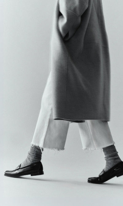 Plümo  Person in long coat, cropped frayed white pants, Alpaca Socks - Slate Grey, and black loafers walking, mid-torso down.