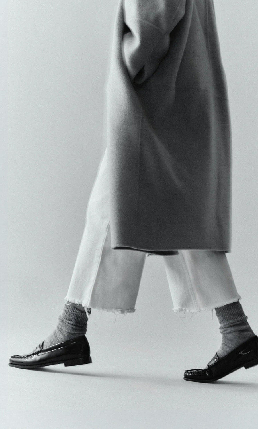 Plümo  Person in long coat, cropped frayed white pants, Alpaca Socks - Slate Grey, and black loafers walking, mid-torso down.
