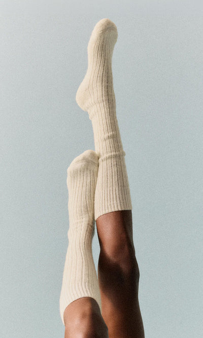 Plümo  Someone wears Clotted Cream Knee Socks with ribbed texture and cushioned sole, legs up against a light backdrop.