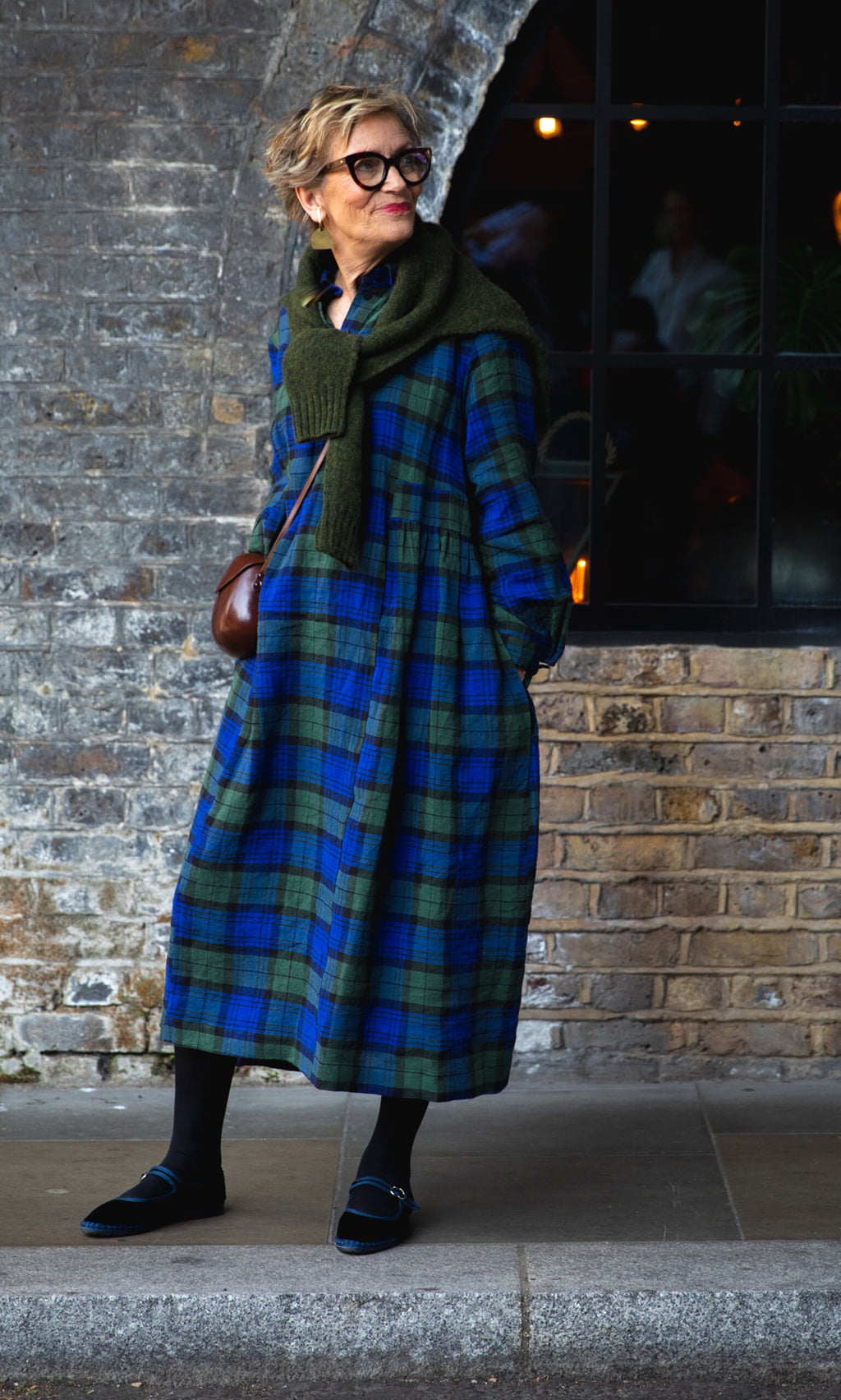 Plümo- Woman in the Laois Tartan Dress with a green sweater and glasses stands by a brick wall, smiling.