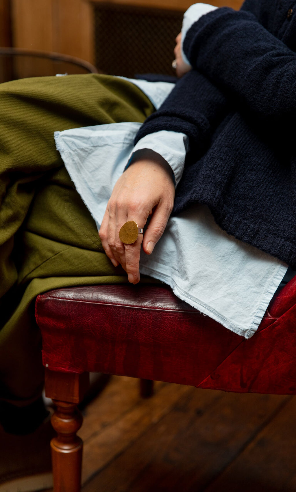 Plümo- A person sits on a red chair, wearing layered blue, white, and green clothes and a handmade Brass Pebble Ring.