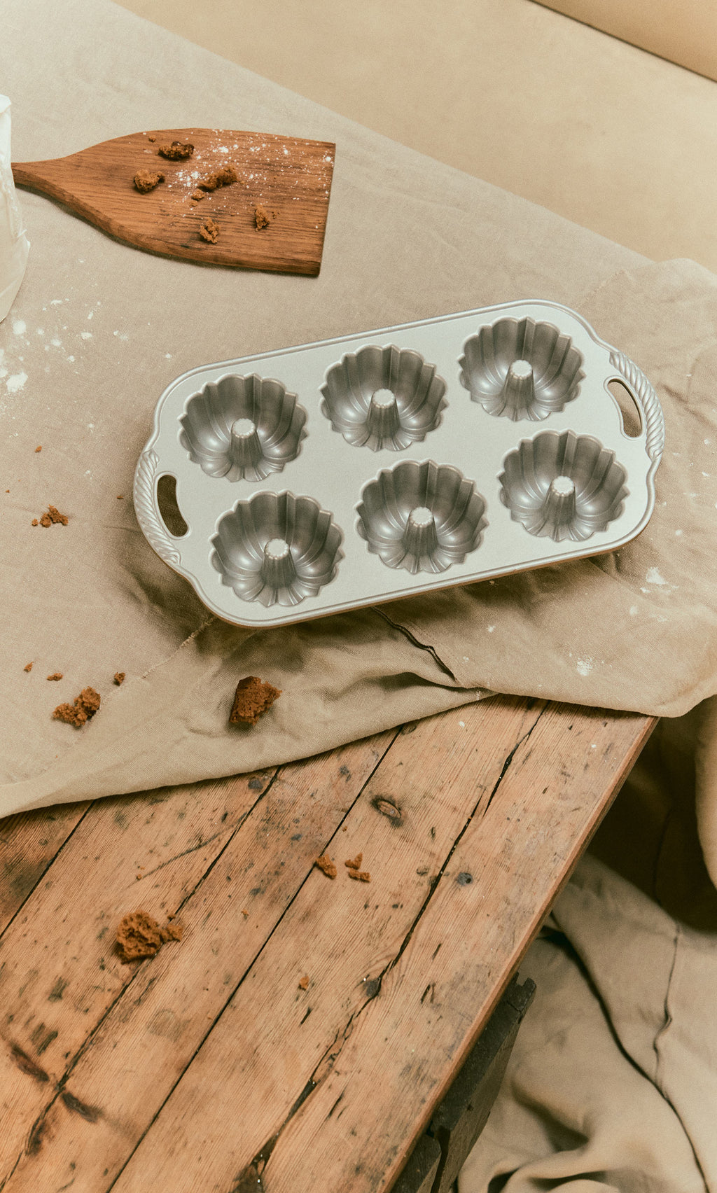 Plümo  A cast aluminium Bundtlette Form with six fluted molds resting on a cloth-covered wooden table.