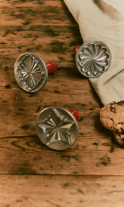 Plümo  Three cast aluminium Cookie Stamps with wooden handles sit on a wooden table next to a stack of cookies.