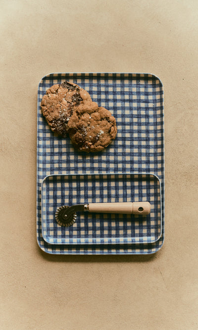 Plümo  A Dough Wheel with a wooden handle rests on a blue and white checkered tray with two cookies.