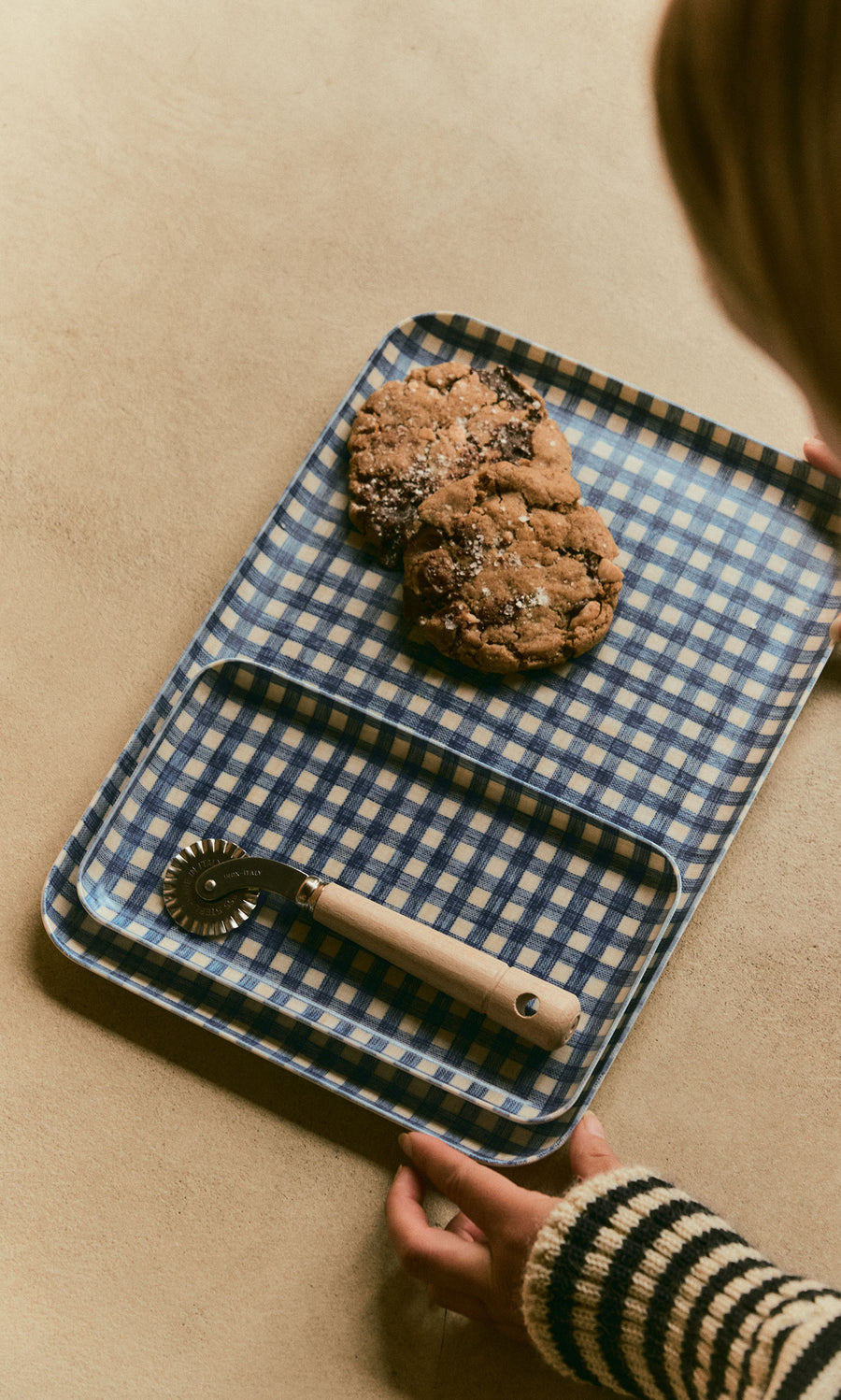Plümo  A Dough Wheel with a wooden handle rests next to two chocolate chip cookies on nested blue checkered trays.