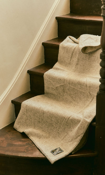 Plümo  The Kilkenny Blanket - Fossil in beige drapes over dark wood stairs, its small label showing at the edge.
