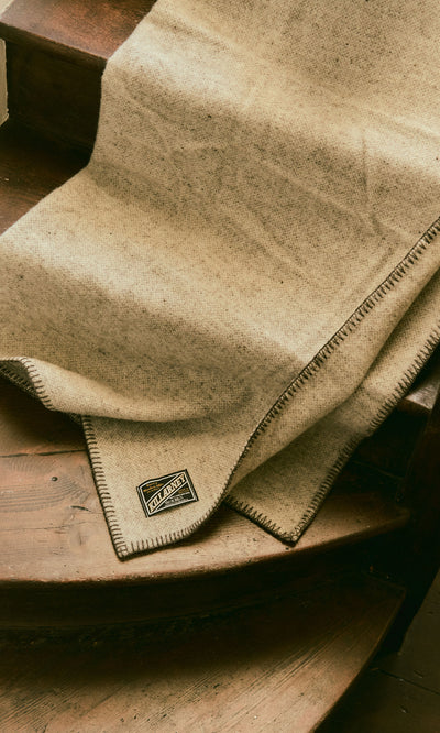 Plümo  Kilkenny Blanket - Fossil, a pure new wool blanket in beige, with label, draped over wooden stairs.