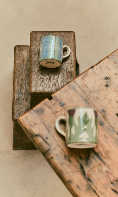 Plümo  Two handmade Blue Tartan Mug pottery mugs with patterned designs sit on stacked wooden stools.