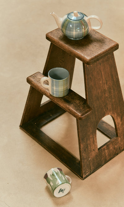 Plümo  A Tartan Teapot with a blue tartan glaze and two patterned mugs resting on and beside a wooden step stool.