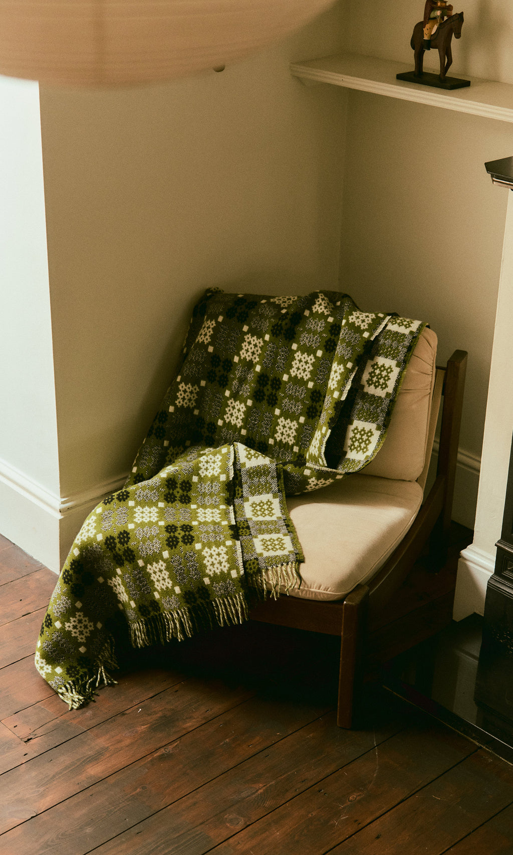 Plümo  A Welsh Blanket - Apple Green is draped over a cushioned chair in the cozy corner of the room.