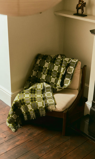 Plümo  A Welsh Blanket - Apple Green is draped over a cushioned chair in the cozy corner of the room.