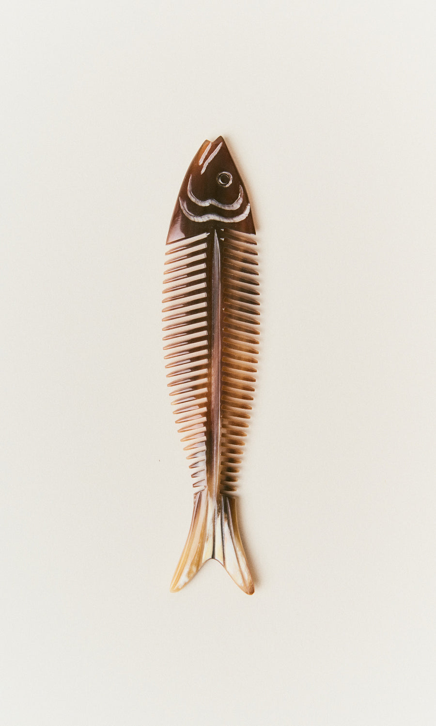 Plümo  The Sardine Comb, brown with white highlights, features a fishbone shape and rests on a plain light background.
