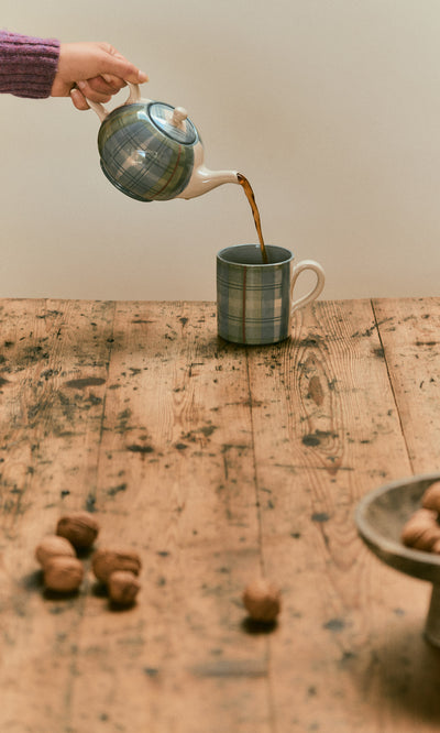 Plümo  A hand pours tea from a blue tartan teapot into a matching Blue Tartan Mug on a wooden table.
