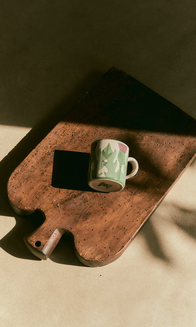 Plümo  A hand-painted Thistle Mug rests on a wooden cutting board, sunlight casting a soft shadow beside it.