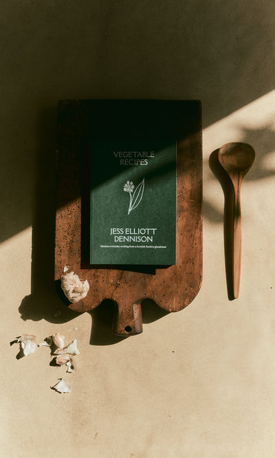 Plümo  Vegetable Recipes By Jess Dennison sits on a wooden cutting board next to a wooden spoon and garlic cloves.