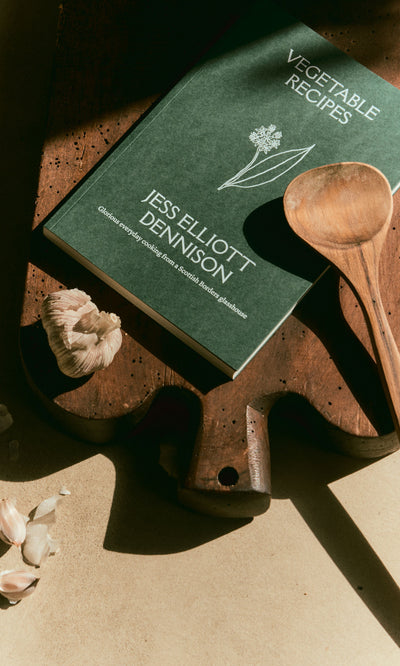 Plümo  Vegetable Recipes By Jess Dennison, a wooden spoon, and garlic sit on a sunlit wooden cutting board.