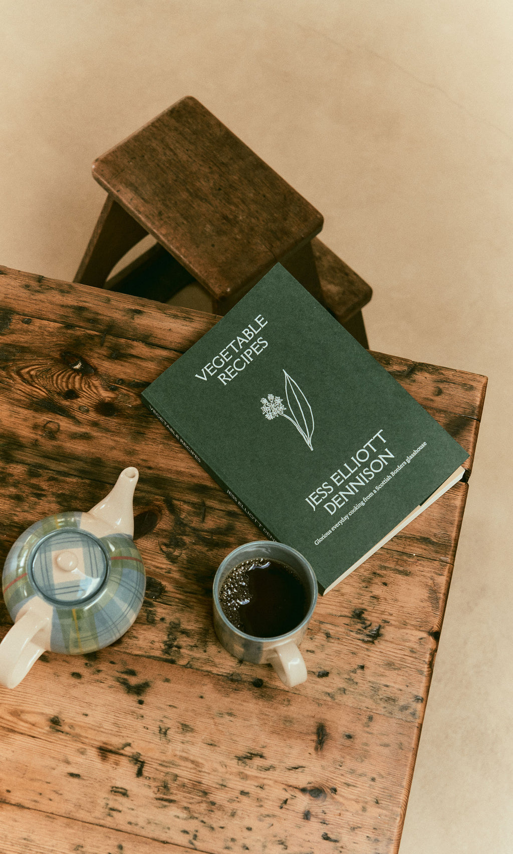 Plümo  Vegetable Recipes By Jess Dennison, a cup of coffee, and teapot sit on a rustic table by a wooden step stool.