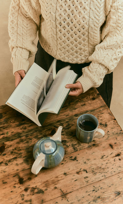 Plümo  Person in a cream sweater reads Vegetable Recipes By Jess Dennison at a rustic table with tea and a teapot.