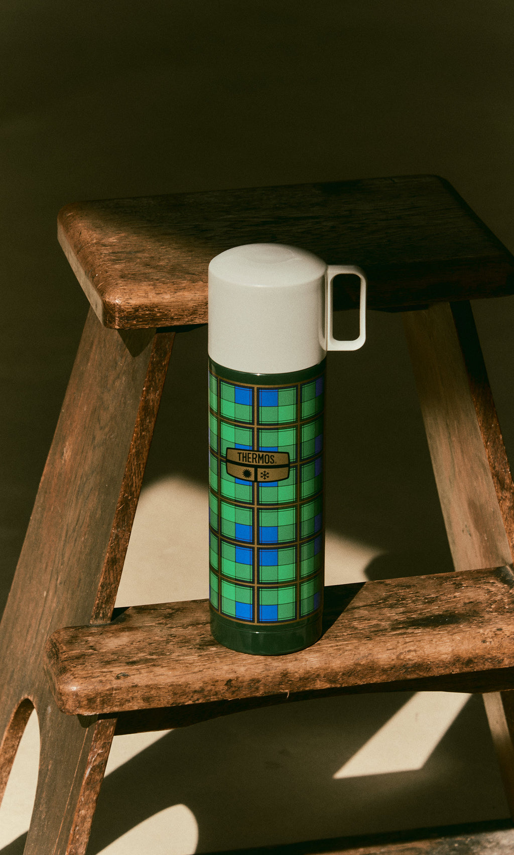 Plümo  Tartan Flask - Green with a white lid, resting on a wooden step stool.