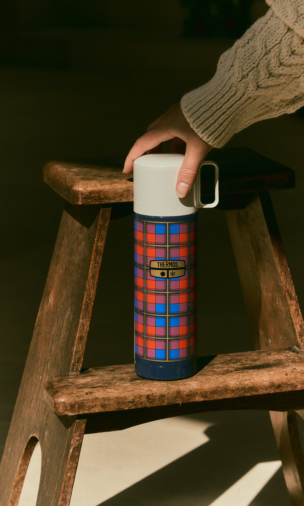 Plümo  A hand holds a blue, red, and yellow Tartan Flask - Blue on a wooden stool.