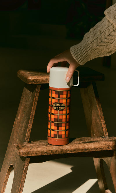Plümo  A hand holds an orange Tartan Flask - Orange with a white lid, resting on a wooden step stool.