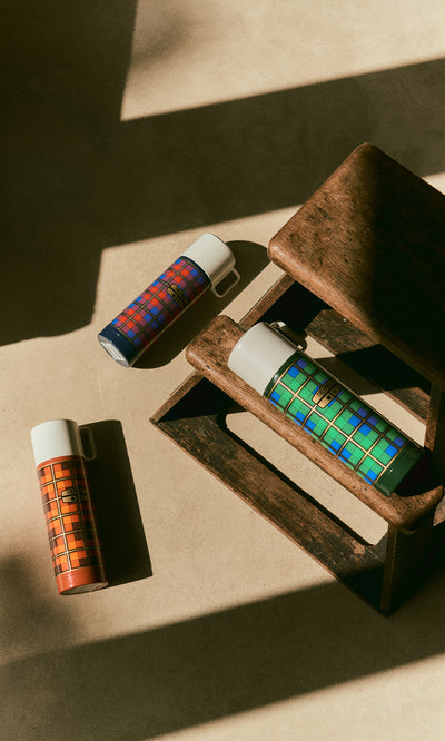 Plümo  Three stainless steel flasks in Tartan Flask - Orange, blue, and green patterns displayed near a wooden step stool.