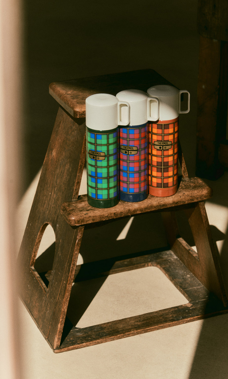 Plümo  Three Tartan Flask - Blue vacuum insulation flasks with white lids sitting on a wooden step stool.