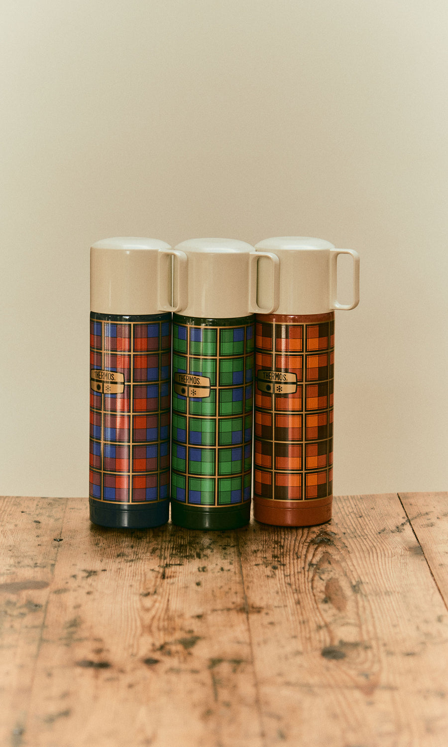 Plümo  Three Tartan Flask - Blue vacuum flasks in blue, green, and orange patterns standing on a wooden surface.