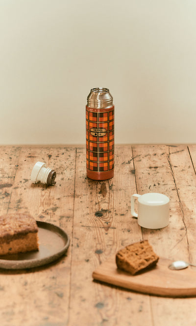 Plümo  Tartan Flask - Orange on a wooden table with a piece of cake and a cup.