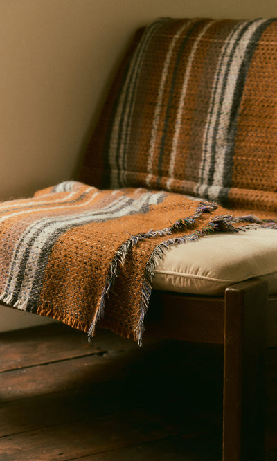 Plümo  A wooden chair with a beige cushion and a Random Throw in orange, gray, and white stripes draped over it.