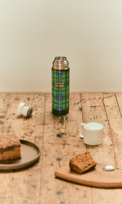 Plümo  A Green Tartan Flask on a wooden table with a white cup, lid, and slices of cake nearby.