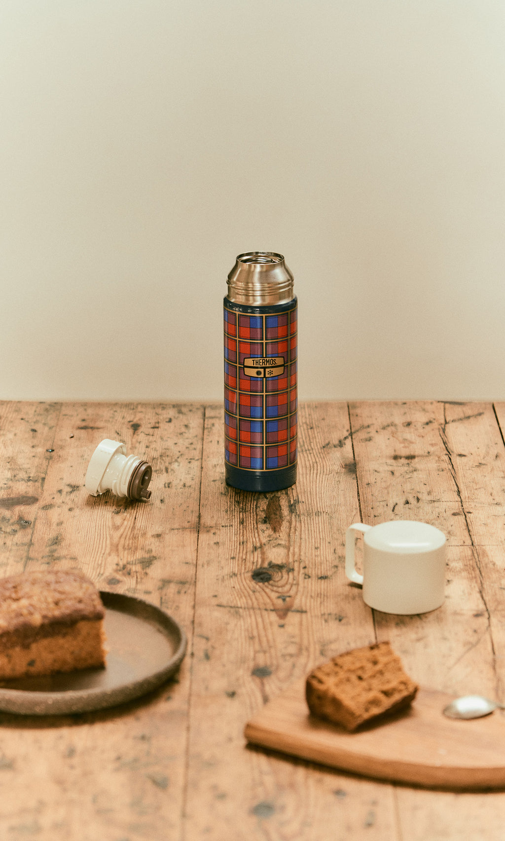 Plümo  A blue, red, and yellow Tartan Flask - Blue sits on a wooden table next to a plate of cake and a small white cup.