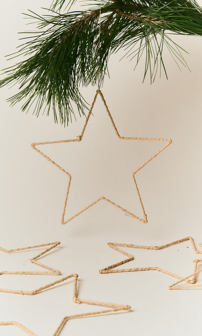 Plümo  A handmade Raffia Christmas Star hangs from a pine branch above two more on a white surface.