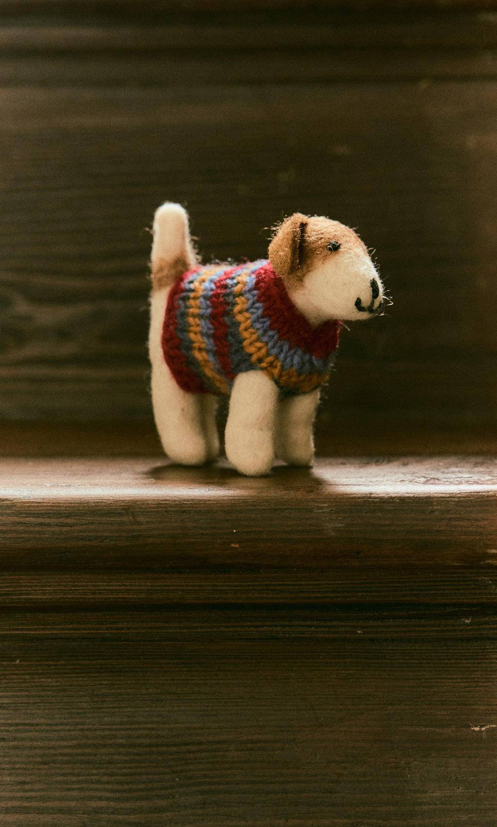 Plümo  Jack in a Jumper, a small hand-felted wool dog in a striped sweater, stands on a wooden staircase.