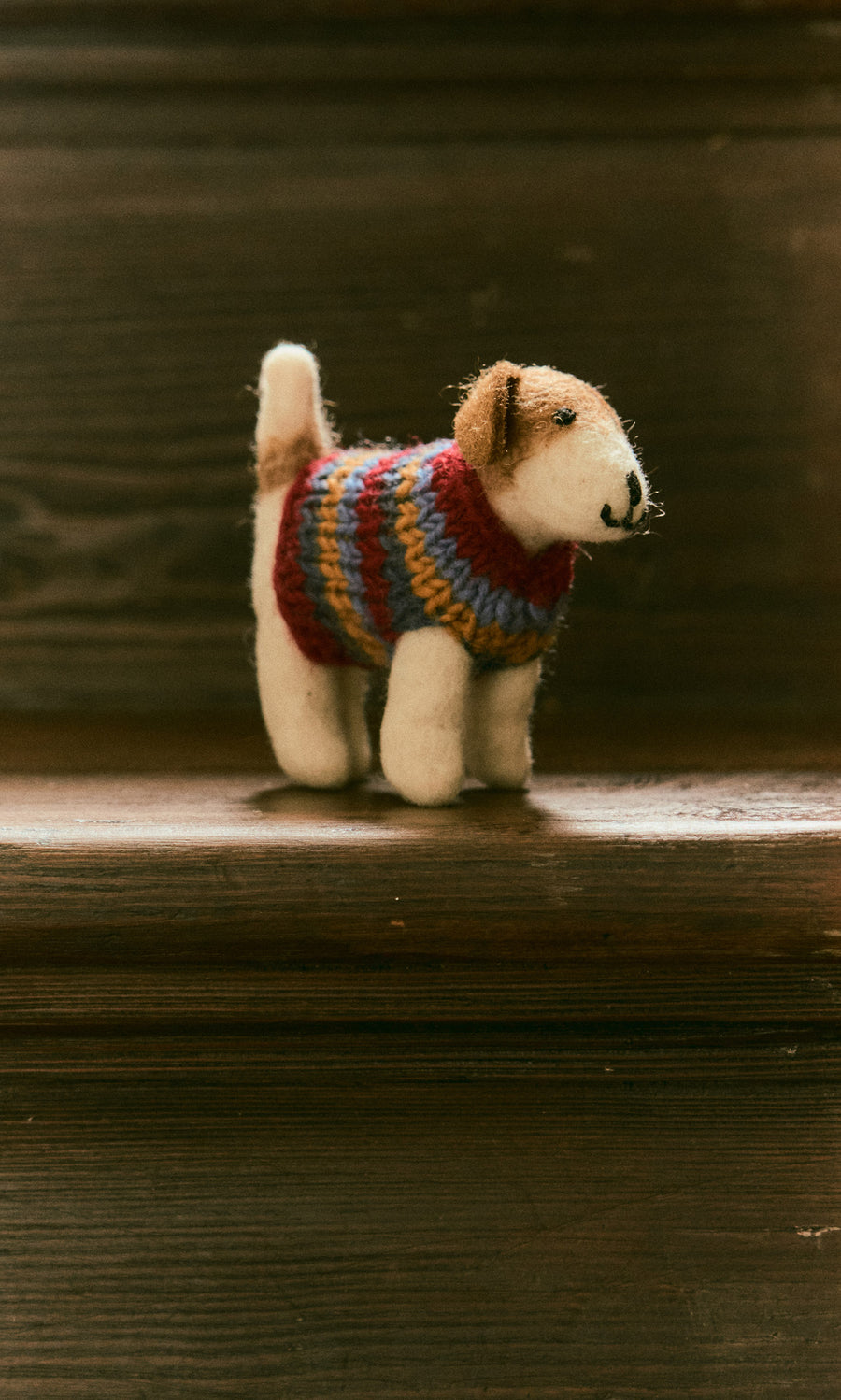 Plümo  Jack in a Jumper, a small hand-felted wool dog in a striped sweater, stands on a wooden staircase.