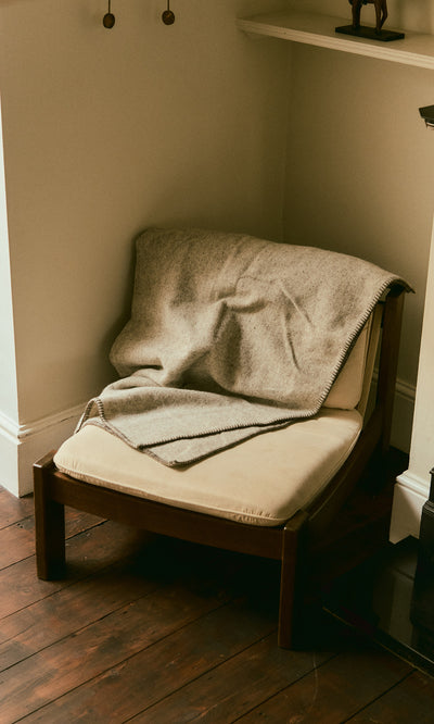 Plümo  A beige chair with the Kilkenny Blanket - Fossil draped over it sits on a wood floor in the corner.