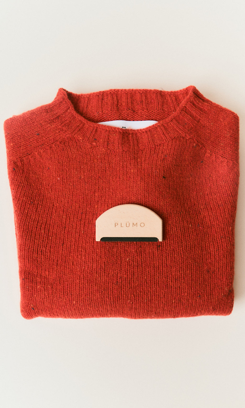Plümo  A folded red sweater with a Wool Comb to remove pilling, set on a light background.