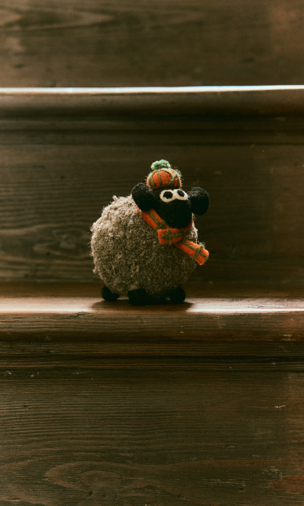 Plümo  Highland Sheep, a plush toy in scarf and hat, handcrafted in Nepal, sits on a wooden stair step.